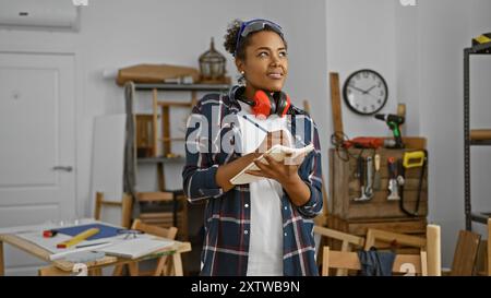 Menuisière féminine réfléchie aux cheveux bouclés prend des notes dans un atelier bien équipé Banque D'Images