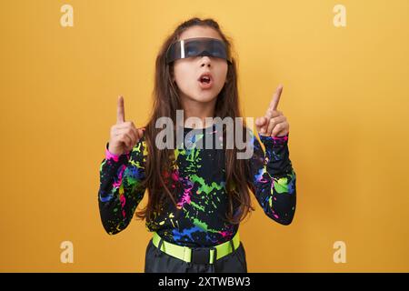 Adorable fille hispanique, stupéfaite dans des lunettes de réalité virtuelle, pointe joyeusement vers le haut. paraissant stupéfaite, elle se tient isolée contre un fond jaune vif Banque D'Images