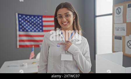 Une jeune femme hispanique souriante avec un autocollant «voté» se tient fièrement dans un centre électoral universitaire avec un drapeau américain. Banque D'Images