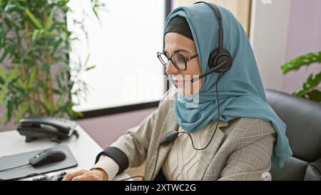 Une femme mature portant le hijab et des lunettes travaille à un casque dans un cadre de bureau lumineux. Banque D'Images