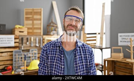 Bel homme barbu dans des lunettes de sécurité se tient dans un atelier de menuiserie bien équipé. Banque D'Images