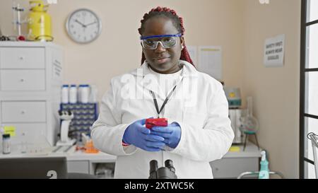 Femme noire avec des tresses portant une blouse de laboratoire et des lunettes de sécurité utilise le téléphone en laboratoire. Banque D'Images