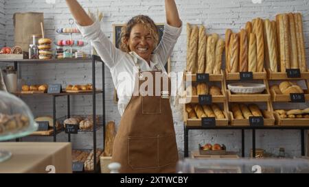 Femme d'âge moyen levant les bras en célébration à l'intérieur d'une boulangerie remplie de divers pains et pâtisseries, mettant en valeur une atmosphère heureuse et accueillante. Banque D'Images