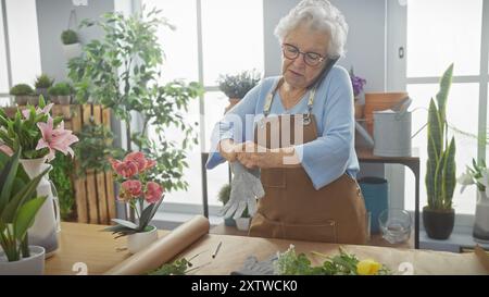 Femme senior portant tablier met des gants dans un magasin de fleurs vibrant tout en parlant au téléphone. Banque D'Images