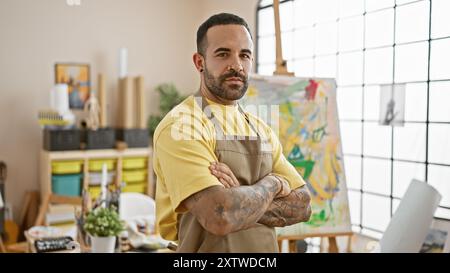 Homme barbu confiant avec des tatouages debout dans un studio d'art, incarnant la créativité et le professionnalisme décontracté. Banque D'Images