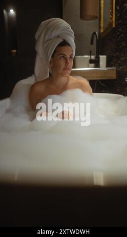 Une jeune femme se détend dans un bain moussant luxueux, ses cheveux enveloppés dans une serviette, dans un décor de salle de bain élégant. Banque D'Images