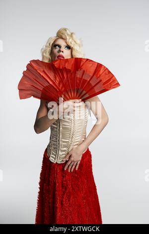 Une drag queen dans une perruque blonde, une jupe rouge et un corset pose dramatiquement, tenant un éventail rouge devant elle. Banque D'Images