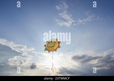 Un ballon souriant en forme de soleil flotte contre un ciel bleu vif avec des nuages moelleux dispersant la lumière du soleil. Banque D'Images