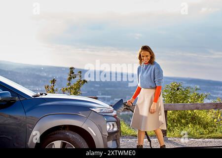 Femme dans une tenue élégante sourit tout en chargeant une voiture électrique avec une vue panoramique en arrière-plan. Banque D'Images