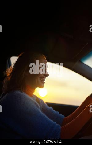 Femme souriant tout en conduisant une voiture au coucher du soleil, avec la lumière chaude du soleil illuminant son visage de côté. Banque D'Images