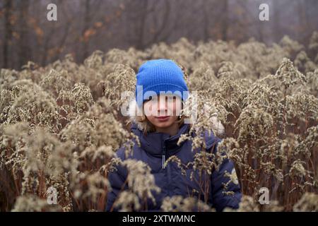 Une jeune personne portant un bonnet bleu et un manteau d'hiver entouré de grandes plantes fanées par une journée brumeuse. Banque D'Images