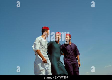 Trois hommes souriants en tenue traditionnelle et des fezzes rouges marchent ensemble sous un ciel bleu clair. Banque D'Images