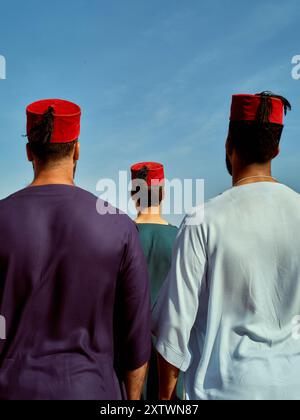 Trois individus portant des chapeaux rouges traditionnels et des vêtements colorés debout contre un ciel bleu clair. Banque D'Images