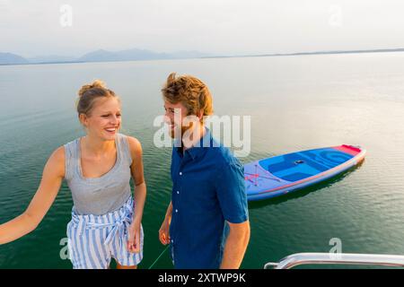 Couple souriant debout sur un quai à côté d'un lac serein avec un bateau bleu flottant à proximité pendant le coucher du soleil. Banque D'Images