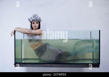 Une jeune femme, ornée d'une couronne chatoyante et d'une queue de sirène, regarde de l'intérieur d'un réservoir rempli d'eau. Banque D'Images