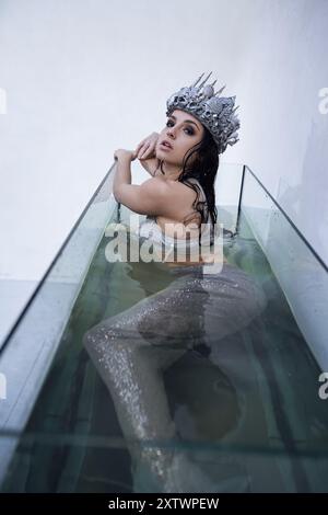 Une jeune femme, habillée en sirène, avec de longs cheveux foncés, est assise dans une enceinte en verre remplie d'eau, portant une couronne d'argent. Banque D'Images