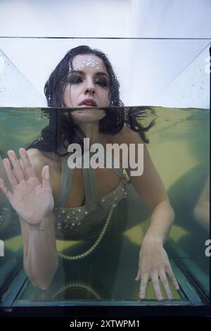 Femme aux cheveux foncés dans le haut chatoyant émerge de l'eau dans le réservoir de verre, regardant triste mais plein d'espoir. Banque D'Images