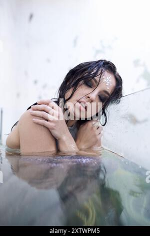 Une jeune femme aux cheveux mouillés, ornée de bijoux chatoyants, est immergée dans une eau claire, regardant de rêve la caméra. Banque D'Images