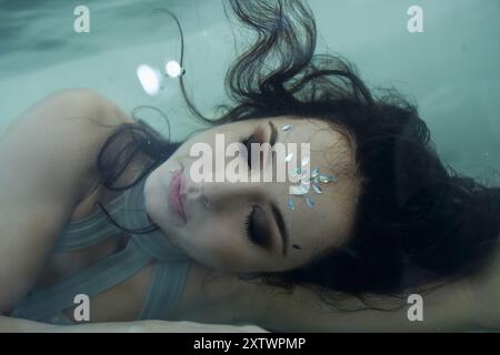 Une jeune femme aux cheveux bruns ornés de bijoux flotte paisiblement dans un aquarium, les yeux fermés dans un état onirique. Banque D'Images