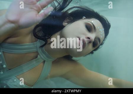 Une jeune femme aux cheveux foncés, ornée de bijoux chatoyants, flotte gracieusement dans un aquarium, les yeux fermés alors qu'elle embrasse l'eau. Banque D'Images