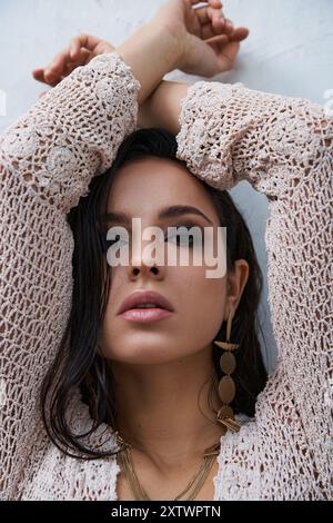 Jeune femme aux cheveux foncés en haut en tricot blanc pose avec les bras croisés au-dessus de la tête, regardant la caméra. Banque D'Images