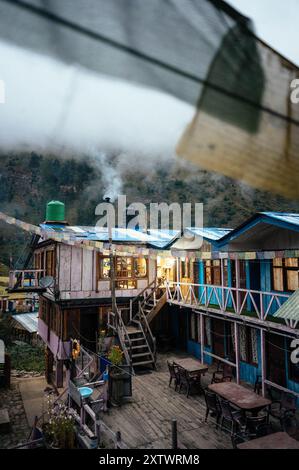 Tôt le matin dans un chalet de montagne rustique avec de la fumée qui s'élève d'une cheminée, des cabanes en bois colorées et des drapeaux de prière flottant dans la brise. Banque D'Images