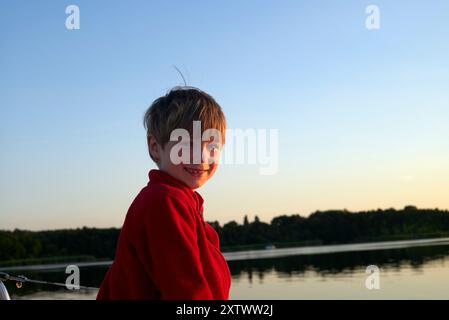 Jeune garçon dans un sweat à capuche rouge souriant par un lac tranquille au coucher du soleil Banque D'Images