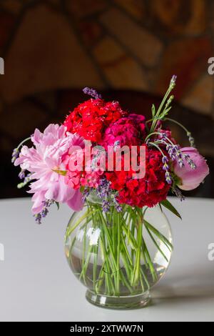 Un bouquet vibrant de pivoines roses, d'hortensias rouges et de lavande violette dans un vase en verre transparent sur une table blanche avec un fond de cheminée chaud et flou. Banque D'Images