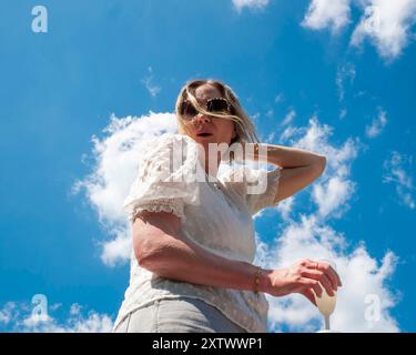 Femme en lunettes de soleil debout contre un ciel bleu clair avec des nuages, paraissant contemplative et tenant un vêtement blanc. Banque D'Images