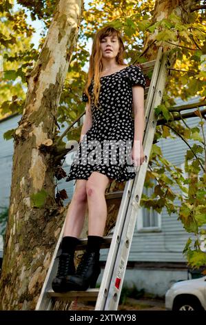 Une femme en robe noire à pois se tient sur une échelle appuyée contre un arbre, avec une expression sereine. Banque D'Images