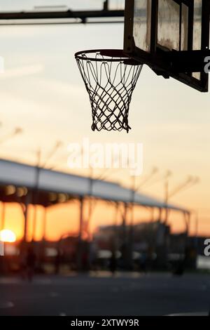 Vue sur le coucher du soleil à travers un panier de basket-ball sur un terrain extérieur avec un paysage urbain flou en arrière-plan. Banque D'Images