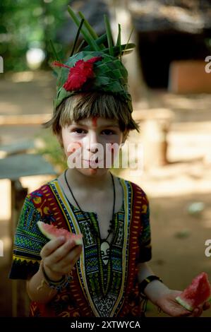 Enfant dans une tenue ethnique vibrante portant une couronne de feuilles et de la peinture de joue tient une tranche de pastèque. Banque D'Images