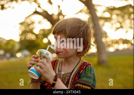 Jeune enfant dans une chemise colorée sirotant une boisson à travers une paille à l'extérieur au coucher du soleil. Banque D'Images