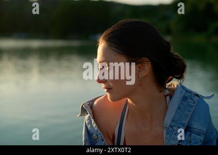 Femme sereine regardant au-dessus d'un lac tranquille au coucher du soleil, avec la lumière chaude du soleil illuminant son profil. Banque D'Images