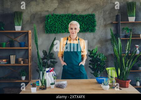 Photo de brillante femme Happy Age habillé tablier rassemblant des plantes à l'intérieur de l'atelier à la maison Banque D'Images