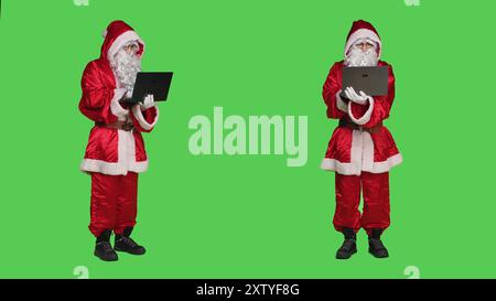 Le Père Noël utilise un ordinateur portable sur l'écran vert, portant un costume de noël festif avec une barbe blanche et naviguant sur le site Web. Modern saint Nick fonctionne sur pc portable, réseau en ligne en studio. Banque D'Images