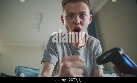 Adolescent caucasien excité joue à un jeu vidéo en ligne sur console à l'aide d'une manette sans fil, est assis sur un canapé. Adolescent garçon se réjouit émotionnellement de la victoire et regarde la caméra. Concept de jeu à domicile. Banque D'Images