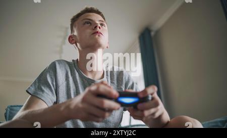 Adolescent caucasien excité joue à un jeu vidéo en ligne sur console à l'aide d'une manette sans fil, est assis sur un canapé. Adolescent garçon se réjouit émotionnellement de la victoire et regarde la caméra. Concept de jeu à domicile. Banque D'Images