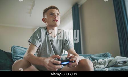 Adolescent caucasien excité joue à un jeu vidéo en ligne sur console à l'aide d'une manette sans fil, est assis sur un canapé. Adolescent garçon se réjouit émotionnellement de la victoire et regarde la caméra. Concept de jeu à domicile. Banque D'Images