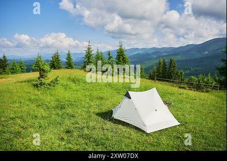 Tente touristique blanche au soleil, au sommet d'une colline herbeuse, entourée de jeunes pins. Vue panoramique sur les montagnes luxuriantes et vallonnées sous un ciel bleu vif avec des nuages dispersés, créant un endroit de camping idyllique. Banque D'Images