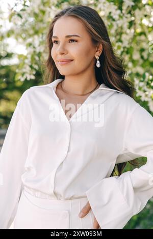 Portrait d'une belle fille aux cheveux longs près d'un arbre en fleurs dans des vêtements blancs tendance Banque D'Images