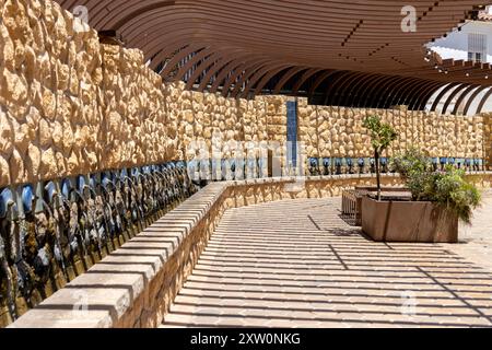 Une longue fontaine murale en pierre sous une pergola en bois voûtée dans un espace public extérieur avec beaucoup de lumière du soleil. Banque D'Images