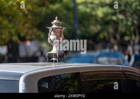 Un samovar décoratif argenté repose au sommet d'une voiture garée dans un quartier animé de la ville. Banque D'Images