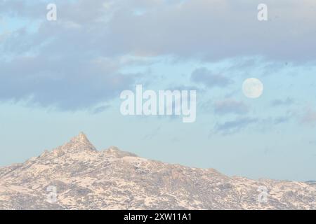 Levez la lune au-dessus d'un pic accidenté et enneigé, entouré de rafales de nuages sur un ciel bleu serein : un moment de tranquillité capturé dans la vaste nature sauvage Banque D'Images