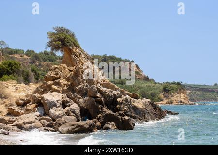 Vagues s'écrasant contre les rochers sur une plage ensoleillée de l'île grecque avec un arbre seul poussant sur une formation rocheuse Banque D'Images
