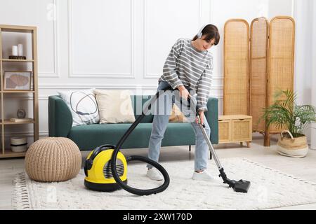 Jeune femme portant des écouteurs nettoyage tapis avec aspirateur dans le salon Banque D'Images