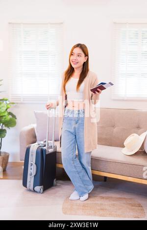 Une jeune femme se tient à côté de sa valise, tenant une carte d'embarquement et un passeport, souriante et prête pour son voyage, avec un canapé confortable et un chapeau à l'arrière Banque D'Images