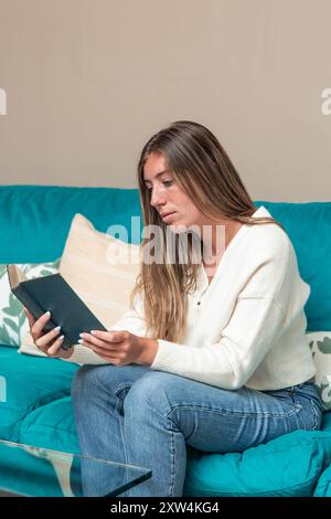 femme blonde verticale est confortablement assise sur son canapé à la maison, profondément engagée dans la lecture d'un livre. La scène transmet un sentiment de relaxation et de tranqui Banque D'Images