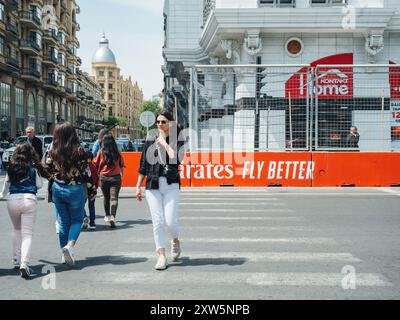 Bakou, Azerbaïdjan - 3 mai 2019 : une élégante femme azérie traverse la rue à Bakou après la course de formule 1, avec la signalisation Emirates Fly Better bien en vue sur les barrières de protection, mêlant sophistication et excitation d'un événement mondial. Banque D'Images