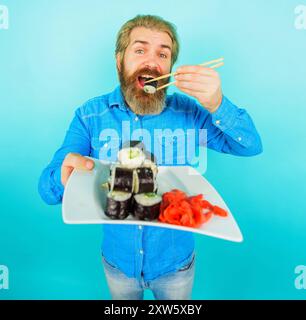 Livraison de sushi ou concept de nourriture à emporter. Homme barbu mangeant un savoureux rouleau de sushi avec des baguettes dégustant de la nourriture japonaise traditionnelle. Homme souriant avec plat Banque D'Images